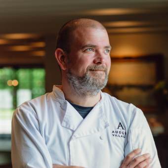 A chef in a white coat stands with arms crossed, gazing to the side. The coat features the "Audley Village" logo. Indoor setting with blurred background.
