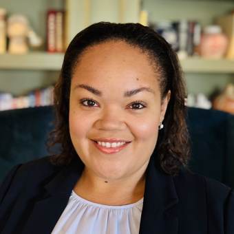 A woman with shoulder-length curly hair, wearing a dark blazer and light blouse, smiles at the camera. A bookshelf with various items is visible in the background.