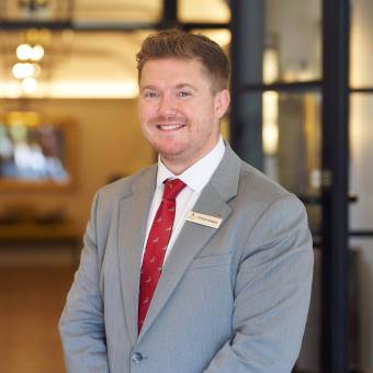 A man in a gray suit and red tie stands indoors, smiling at the camera, wearing a name badge.