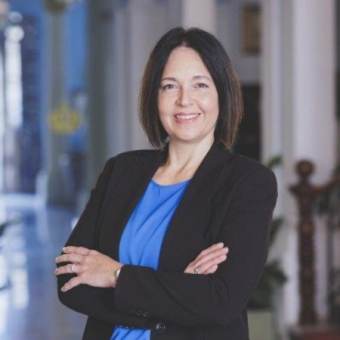 A woman with shoulder-length dark hair wearing a black blazer and blue top with her arms crossed.