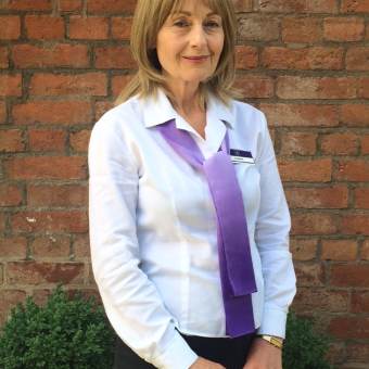A woman with blonde hair in a white shirt and purple scarf stands in front of a brick wall, with potted green plants on either side.