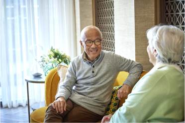 Two elderly people sitting on a yellow sofa, engaged in conversation, with light shining through sheer curtains in the background.