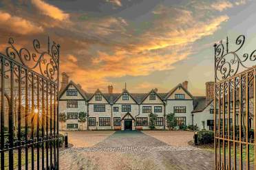 A large Tudor-style mansion with multiple chimneys is seen through open wrought iron gates at sunset, with a cobblestone driveway leading to the entrance.