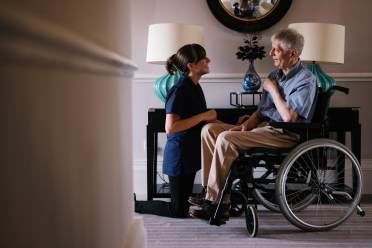 A nurse kneels beside an elderly man in a wheelchair, smiling and engaging in conversation in a well-lit room with lamps and a mirror.