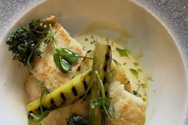 Pan-seared white fish served with grilled leeks, crispy kale, and a light herb sauce in a shallow bowl.
