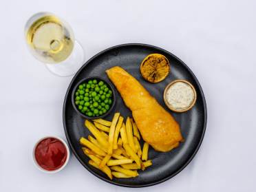Fish and chips on a black plate with a glass of white wine on the side
