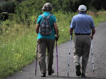 Two people using walking poles on a gravel path. The person on the left has a multicolored backpack, and the person on the right wears a cap. Greenery surrounds the path.