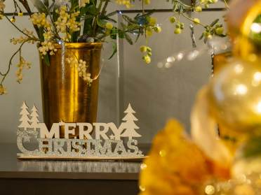A glittery "Merry Christmas" sign sits on a surface next to a gold vase with festive greenery; blurred gold decorations appear in the foreground.