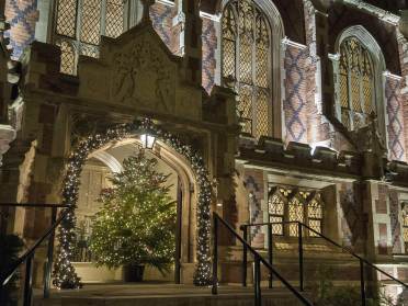 A decorated Christmas tree with lights stands under an arched entrance of a building with illuminated windows at night.