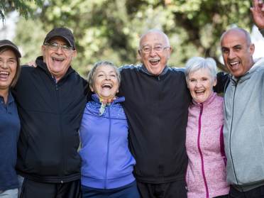 A group of six smiling older adults stand outdoors, wearing casual jackets. Trees are blurred in the background.