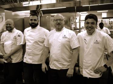 Four chefs in white uniforms stand in a commercial kitchen, smiling.