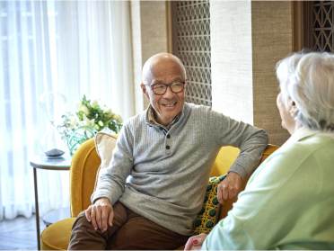 Two elderly people sitting on a yellow sofa, engaged in conversation, with light shining through sheer curtains in the background.