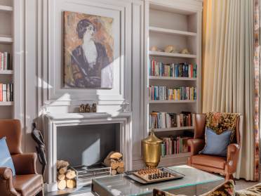 Elegant living room with two armchairs, a fireplace, and built-in bookshelves. A painting hangs above the fireplace, and a chess set is on a glass coffee table in the center.