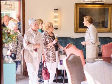 A group of older adults enter a room, one holding a drink and a shopping bag, while a woman in a blazer addresses them. The room features a framed painting and upholstered chairs.