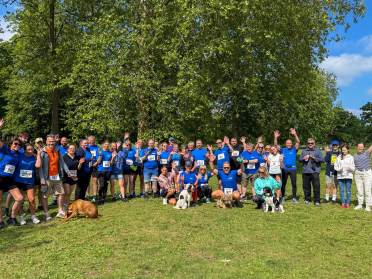 A large group of people, some with dogs, stand on grass in front of trees, wearing athletic clothes and race bibs, smiling and waving at the camera.