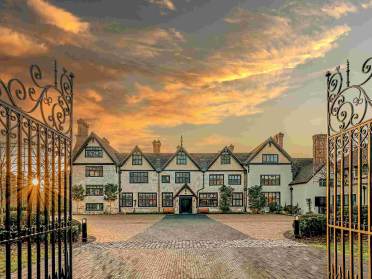 A large Tudor-style mansion with multiple chimneys is seen through open wrought iron gates at sunset, with a cobblestone driveway leading to the entrance.