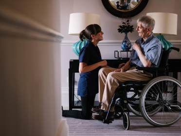 A nurse kneels beside an elderly man in a wheelchair, smiling and engaging in conversation in a well-lit room with lamps and a mirror.