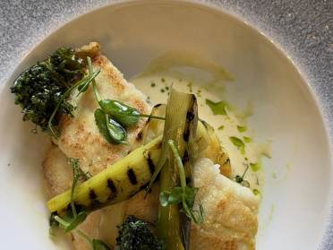 Pan-seared white fish served with grilled leeks, crispy kale, and a light herb sauce in a shallow bowl.