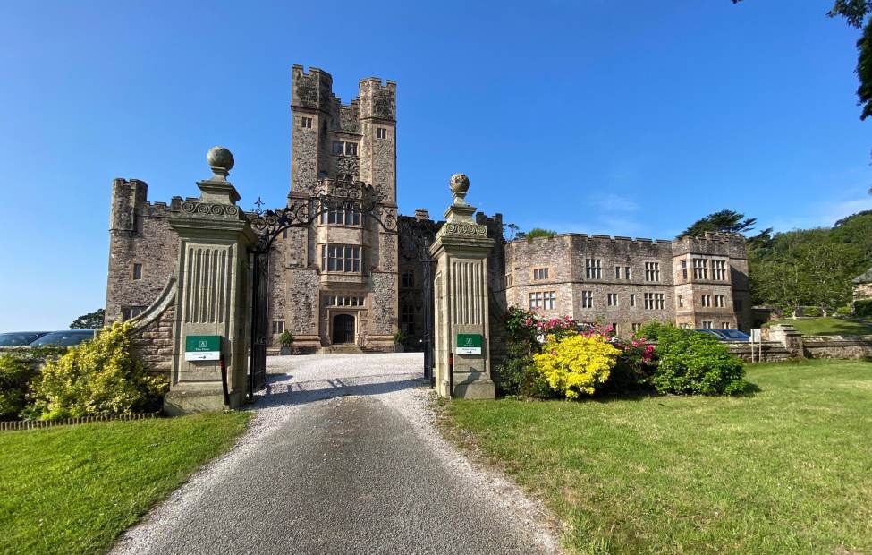 Flete House A historic stone castle with two tall towers, an arched entrance, and a gravel driveway, surrounded by manicured gardens and lawns under a clear blue sky.