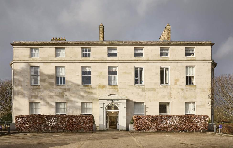 Mote House A large, three-story, historic stone building with symmetrical windows and a central entrance. A hedge surrounds the base, and bare trees are visible in the background.