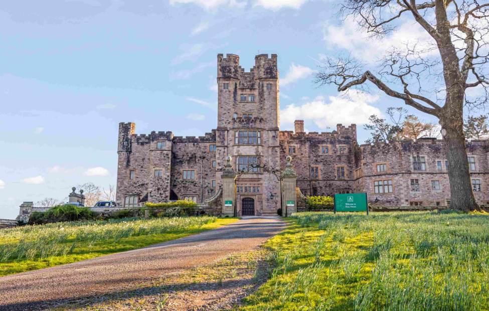 Flete House A stone castle with towers and battlements stands at the end of a gravel path, surrounded by grass and trees under a partly cloudy sky.
