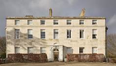 A large, three-story, historic stone building with symmetrical windows and a central entrance. A hedge surrounds the base, and bare trees are visible in the background.
