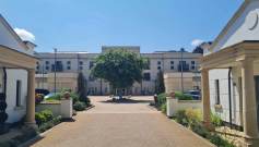 A wide pathway flanked by two white buildings leads to a large central building with balconies, with a leafy tree in the courtyard under a clear blue sky.